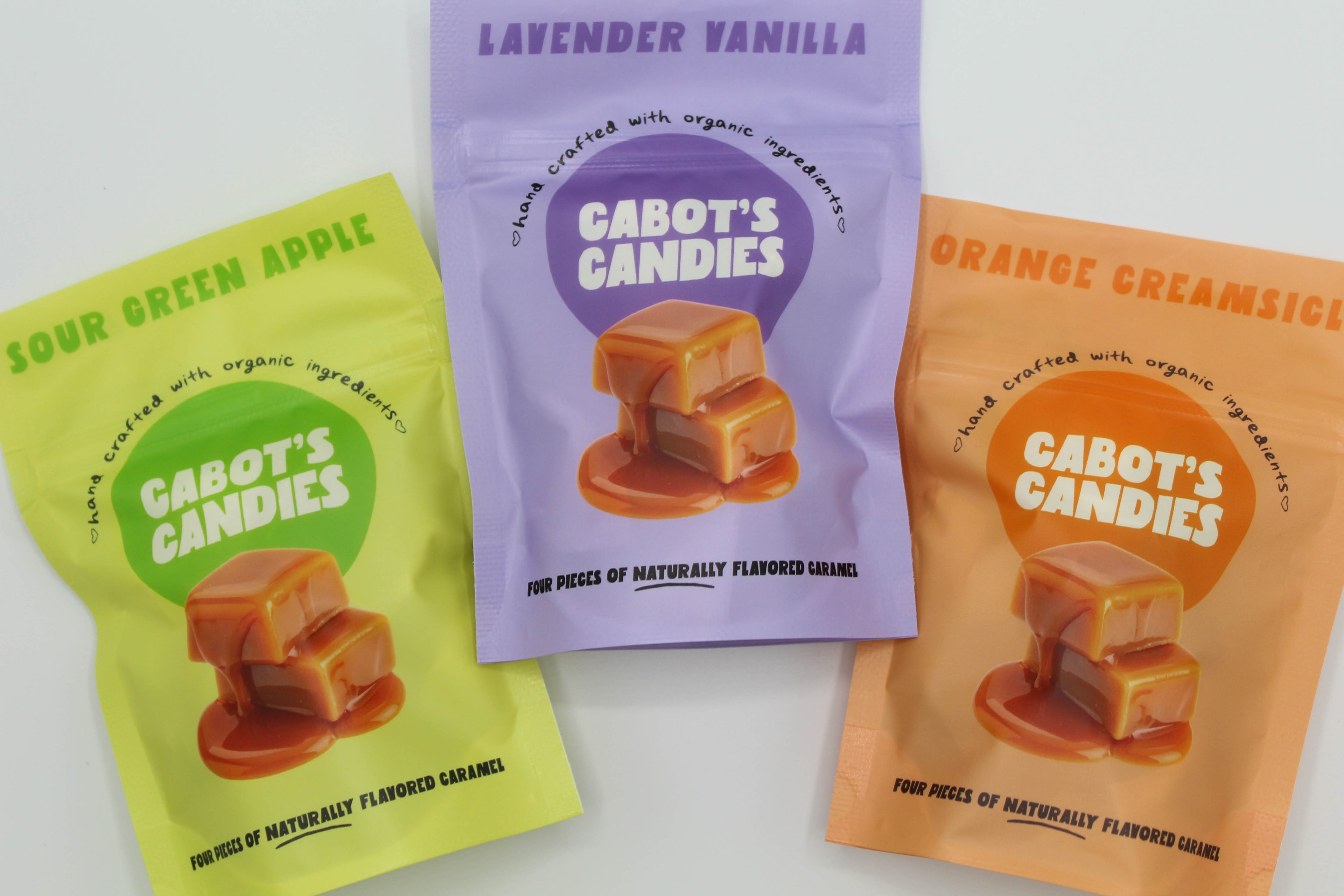Three pouches of flavored caramels sit against a white background. One is flavored sour green apple, one is lavender vanilla, and one is orange creamsicle.