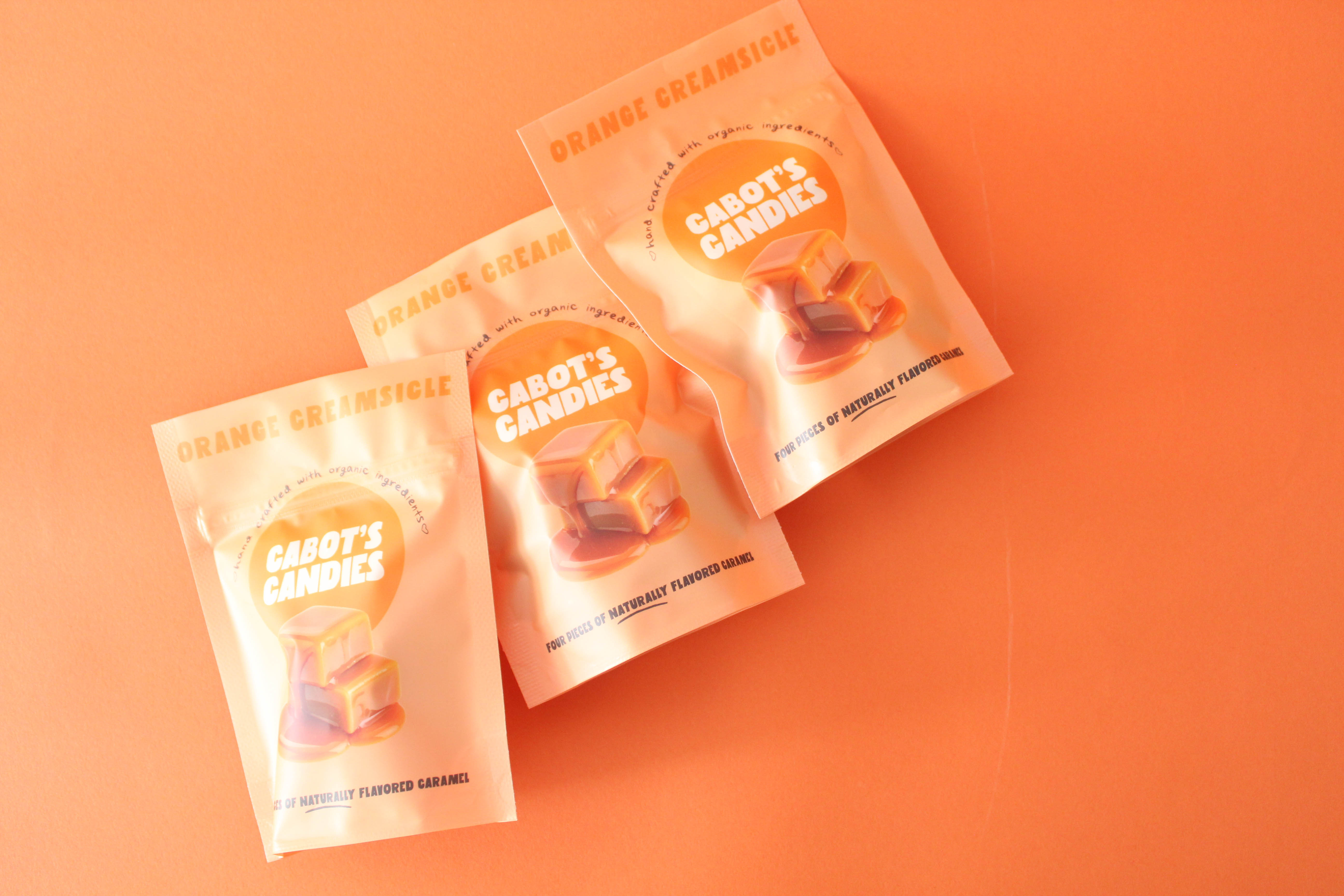 Three pouches of Cabot's Candies Orange Creamsicle flavor arranged diagonally on a solid orange background.