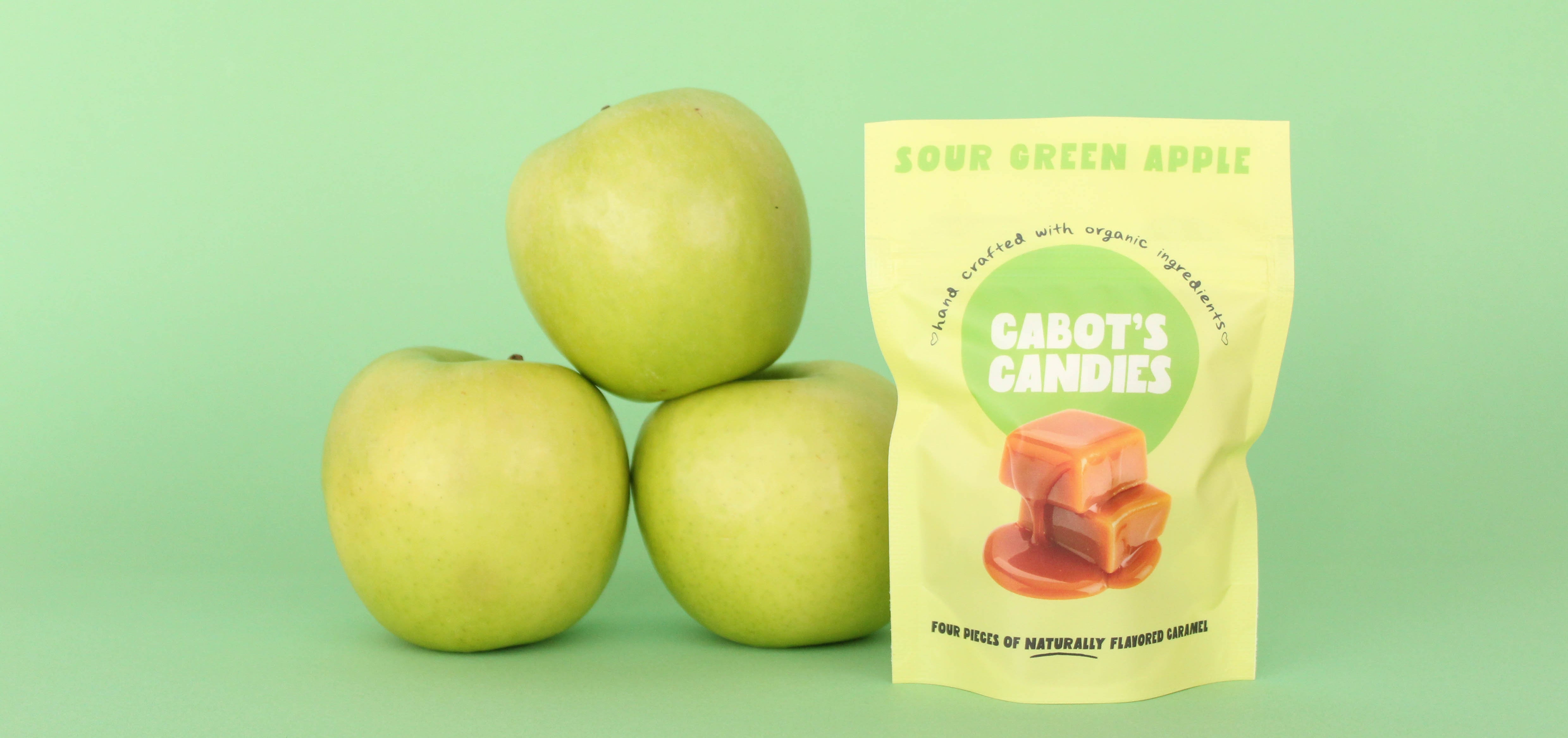 A stand-up pouch of Cabot's Candies Sour Green Apple flavor stands on a light green background next to three whole green apples. The pouch is pale yellow with green and red graphics, and the apples are bright green.